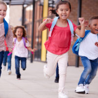 A group of smiling multi-ethnic school kids running in a walkway outside their infant school building after a lesson, close up