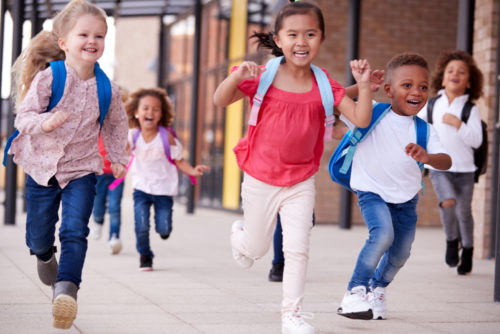 A group of smiling school kids running in a walkway outside a building.