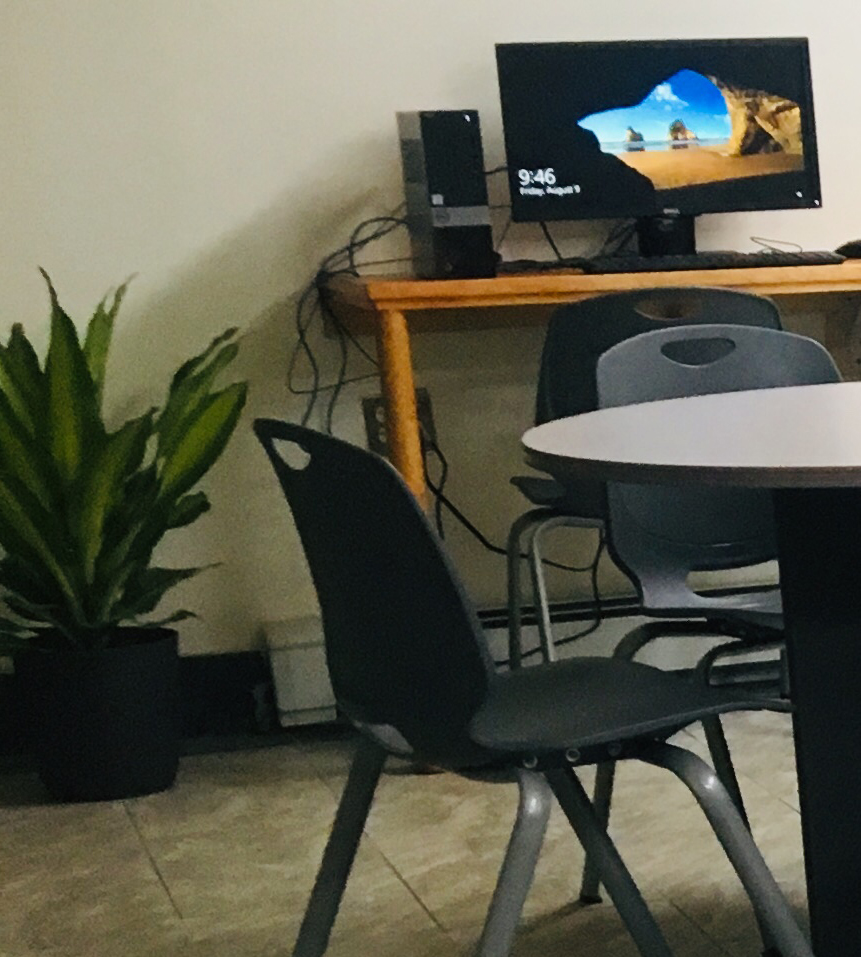 Computer lab at Rico's Place: coffee tables with chairs in foreground, computer and house plant along the wall.